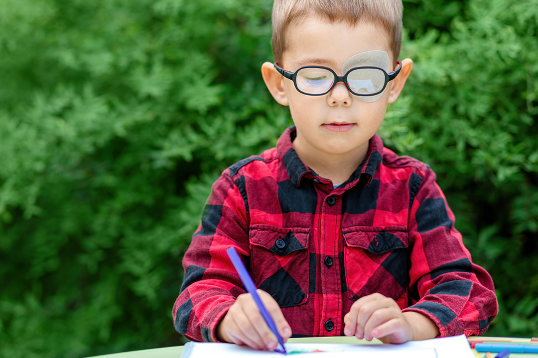 Ein kleiner Junge mit Augenpflaster am linken Auge und Brille auf, malt ein Bild im Garten.