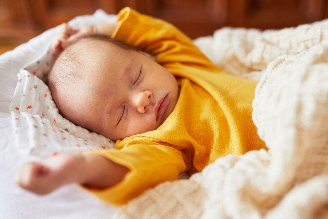 Ein schlafendes Baby in einem gelben Obertail das zugedeckt da liegt.