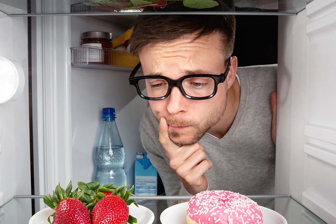 Mann guckt fragend in einen Kühlschrank mit Erdbeeren und einem Donut