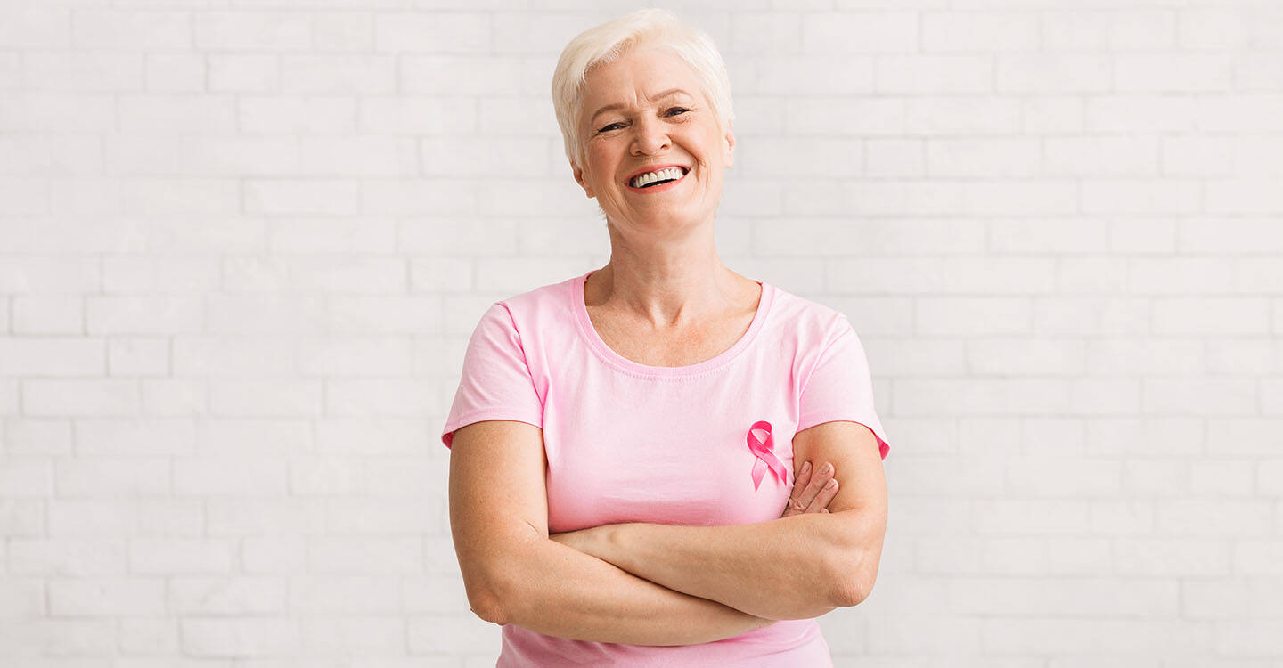 Eine Frau lächelt in die Kamera, dabei trägt sie ein Rosa T-Shirt mit der Brustkrebst Schleife darauf.