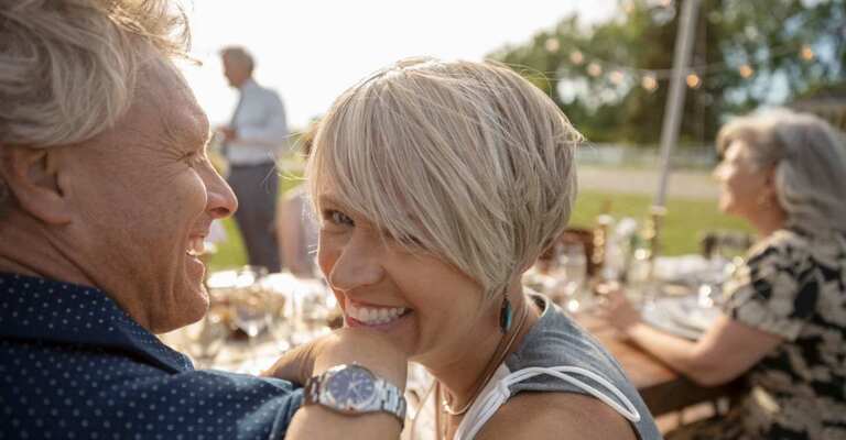 Eine Frau lächelt auf einer Sommerparty über die Schulter ihres Mannes in die Kamera 