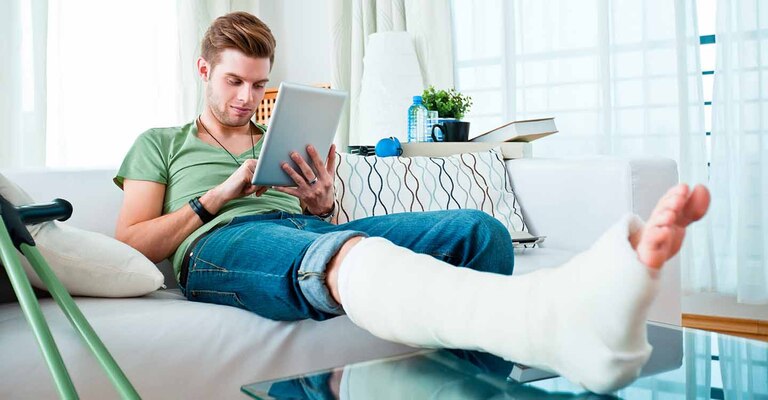 Junger Mann sitzt in einem hellen Raum, mit einem gebrochenen rechten Bein in Gips auf dem Sofa und bedient ein Tablet