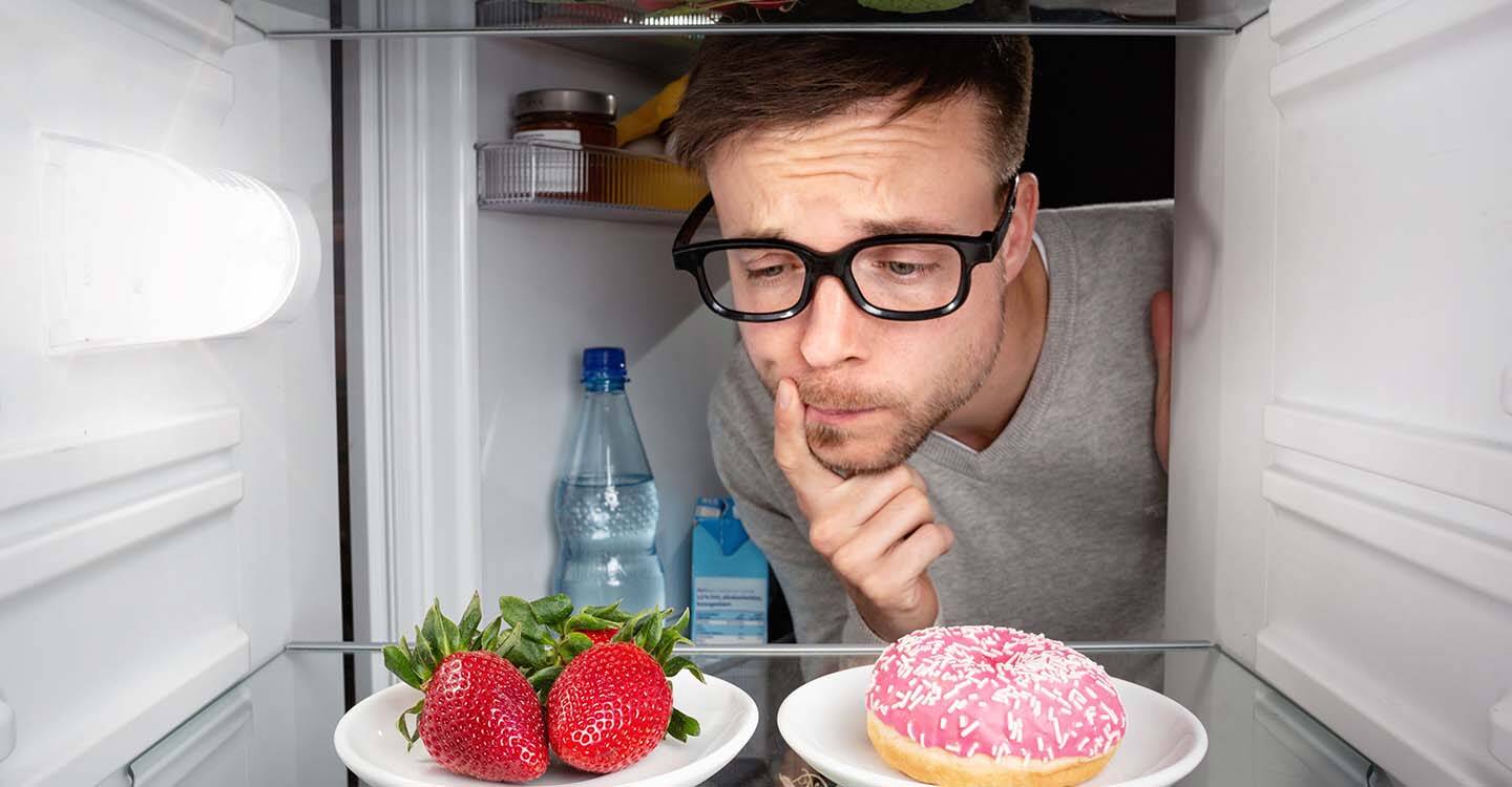 Mann guckt fragend in einen Kühlschrank mit Erdbeeren und einem Donut