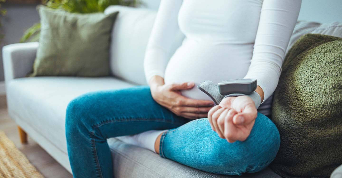 Schwangere Frau sitzt mit einem Blutdruckmessgerät auf dem Sofa