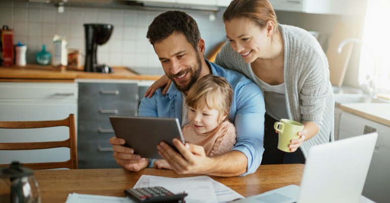 Vater sitz mit Tochter auf dem Schloß am Esstisch und schaut ins Tablet. Ehefrau schaut dabei über die Schulter.