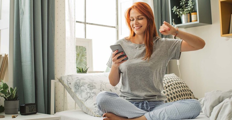 Frau sitzt lachend auf dem Bett und guckt auf ihr Handy