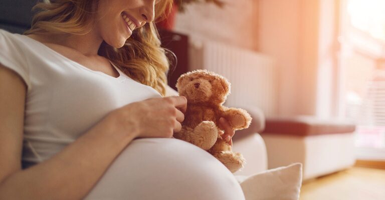 Schwangere Frau sitz auf dem Sofa mit einem Stoffteddy in der Hand.